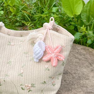 Peut inclure: Un sac fourre-tout beige avec une texture tissée et des broderies florales. Le sac est orné d'un coquillage crocheté blanc et d'un charm en forme d'étoile de mer rose avec des perles. Décoré d'un pompon et d'un cordon rose.