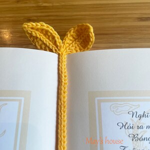 May include: A yellow crocheted bookmark with two leaf-shaped ends. The bookmark is on a white page with text in Vietnamese.