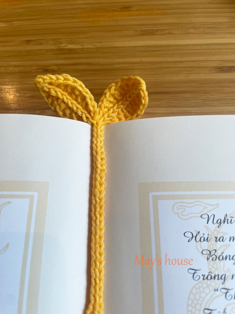 May include: A yellow crocheted bookmark with two leaf-shaped ends. The bookmark is on a white page with text in Vietnamese.