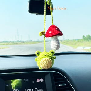 Puede incluir: Amuleto de coche de ganchillo con una rana verde con vientre color crema y una seta roja con lunares blancos. El amuleto cuelga de una cuerda verde, con el texto "May's house" visible.