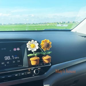 May include: Two crochet flower car air vent decorations. One is a white daisy with a yellow center, and the other is a yellow sunflower. Both are in small brown crochet pots. The car dashboard also displays the text "May's house".