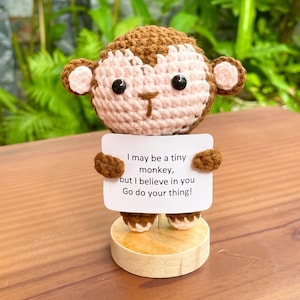 May include: A crocheted monkey figurine with brown and light pink yarn, featuring black bead eyes and a small brown nose. The monkey holds a white rectangular sign that reads: I may be a tiny monkey, but I believe in you Go do your thing!