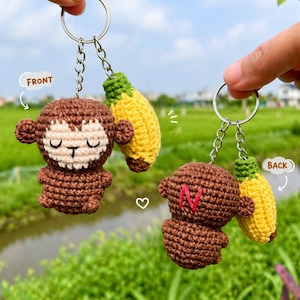 Pode incluir: Dois chaveiros de macaco de crochê feitos à mão com pingentes de banana. Os macacos são marrons com rostos creme e olhos fechados. Um chaveiro tem a palavra "FRONT" e o outro a palavra "BACK". O texto "May's house" está no fundo.