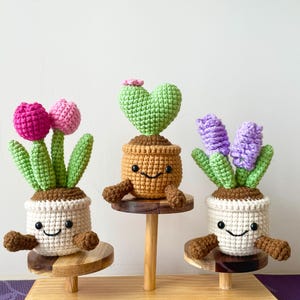 May include: Three charming crochet plant figures with smiling faces. One has pink tulips, another a green heart, and the last has purple flowers. Each plant sits in a white or brown pot with brown arms, displayed on a wooden stand.