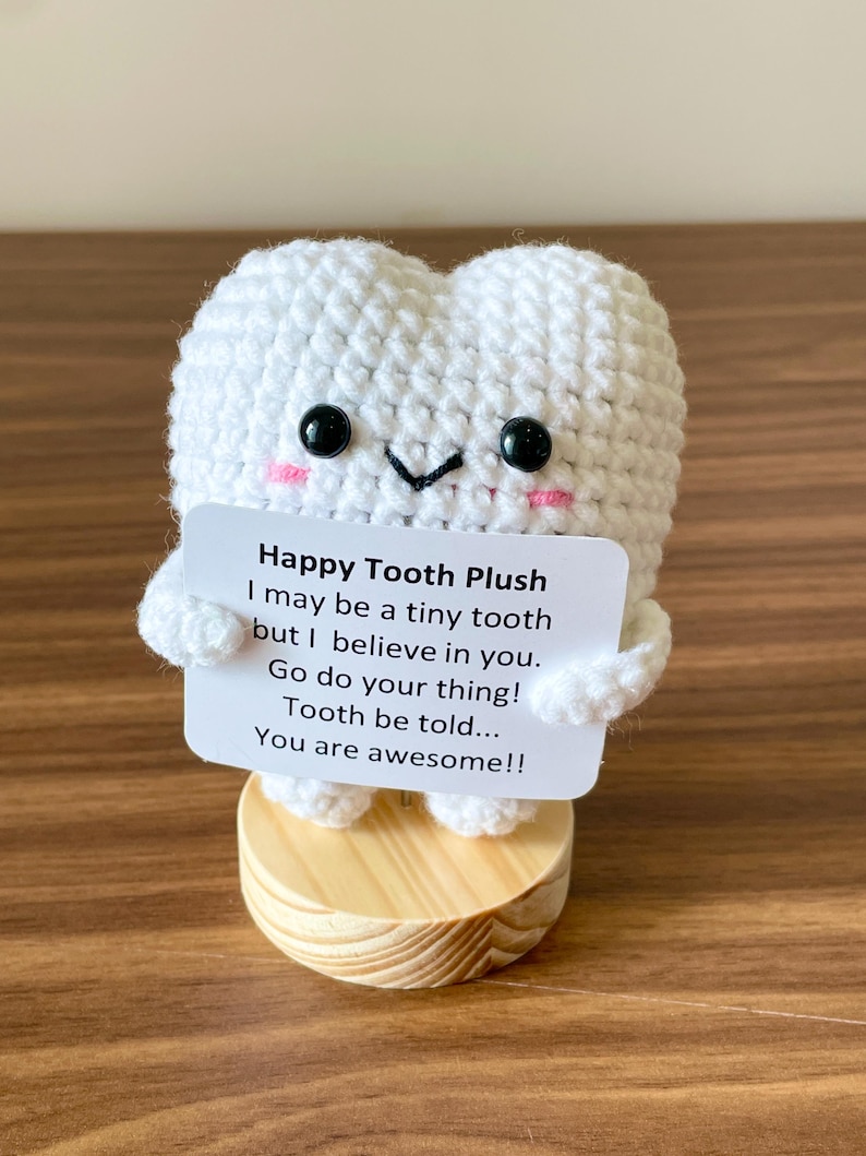 May include: A white crocheted tooth plush with black eyes and pink cheeks, holding a sign that reads "Happy Tooth Plush." The plush is on a small wooden base. The sign has encouraging words.