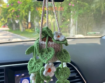 May include: A crocheted hanging plant with green leaves and white flowers. The plant is hanging from a rearview mirror in a car. The plant is a handmade item and has the text "MAY'S HOUSE" on the tag.
