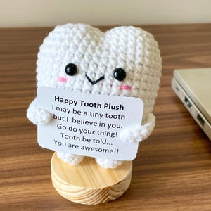 May include: A white crocheted tooth plushie with black eyes and pink cheeks, holding a card that reads "Happy Tooth Plush." The plushie is on a small wooden base. The card has an inspirational message.