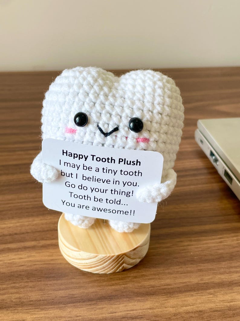 May include: A white crocheted tooth plushie with black eyes and pink cheeks, holding a card that reads "Happy Tooth Plush." The plushie is on a small wooden base. The card has an inspirational message.