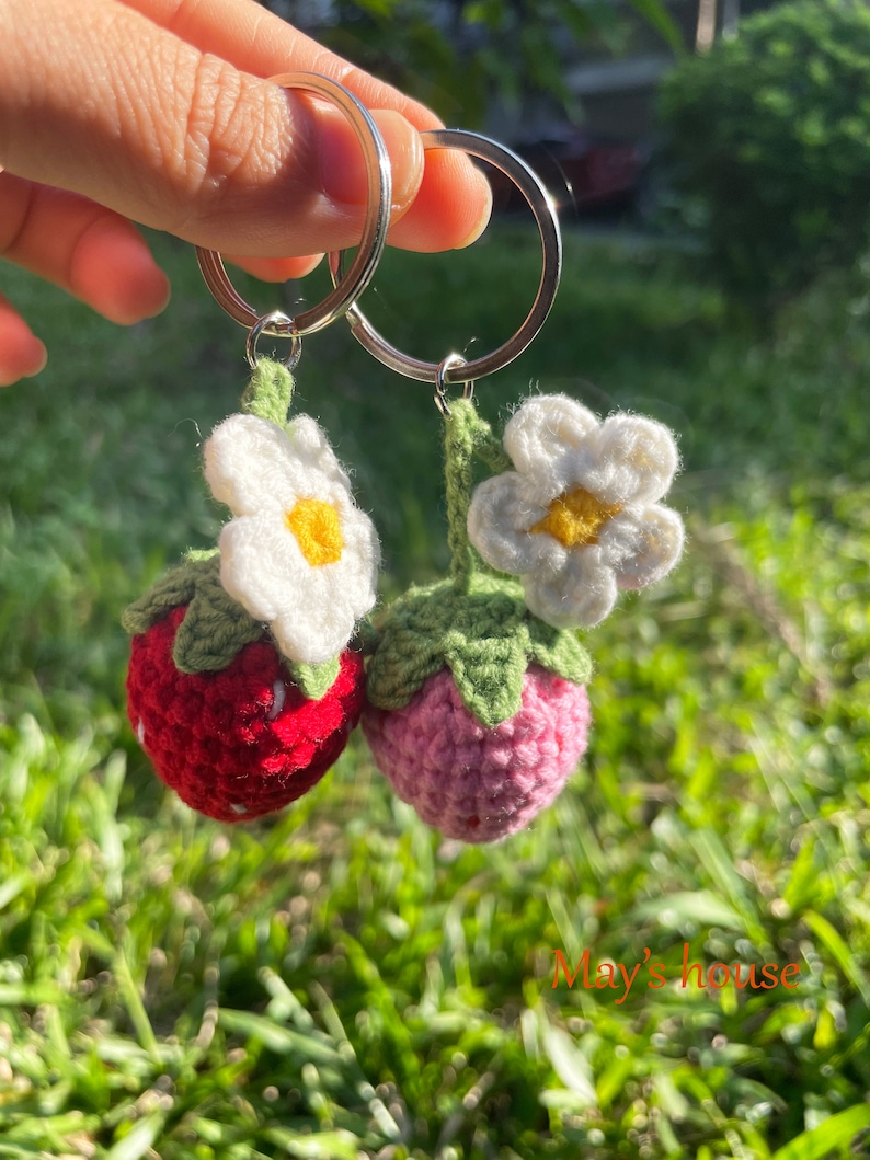 May include: Two crocheted strawberry keyrings with white flowers and green leaves. The keyrings are attached to silver key rings.
