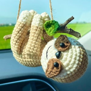 Puede incluir: Un adorno de coche de perezoso hecho a mano con ganchillo. El perezoso es beige y blanco, con detalles marrones para los ojos y la nariz. Cuelga de una cuerda de hilo natural, con una pequeña hoja verde y una rama de madera.