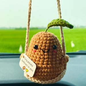 Puede incluir: Adorno de patata marrón tejido a ganchillo, con una hoja verde y una cara sonriente. Cuelga de una cuerda beige y tiene una etiqueta que dice "Tiny potato believes in you".