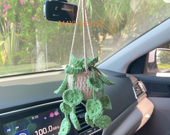 May include: A crocheted hanging plant with green leaves and a cream-colored pot. The plant is hanging from the rearview mirror of a car. The car's radio display shows "100.0 MHz".
