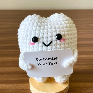 May include: A handmade white crochet tooth with black button eyes, pink cheeks, and a smiling mouth. The tooth holds a small white sign that reads "Customize Your Text." It is mounted on a small wooden base.
