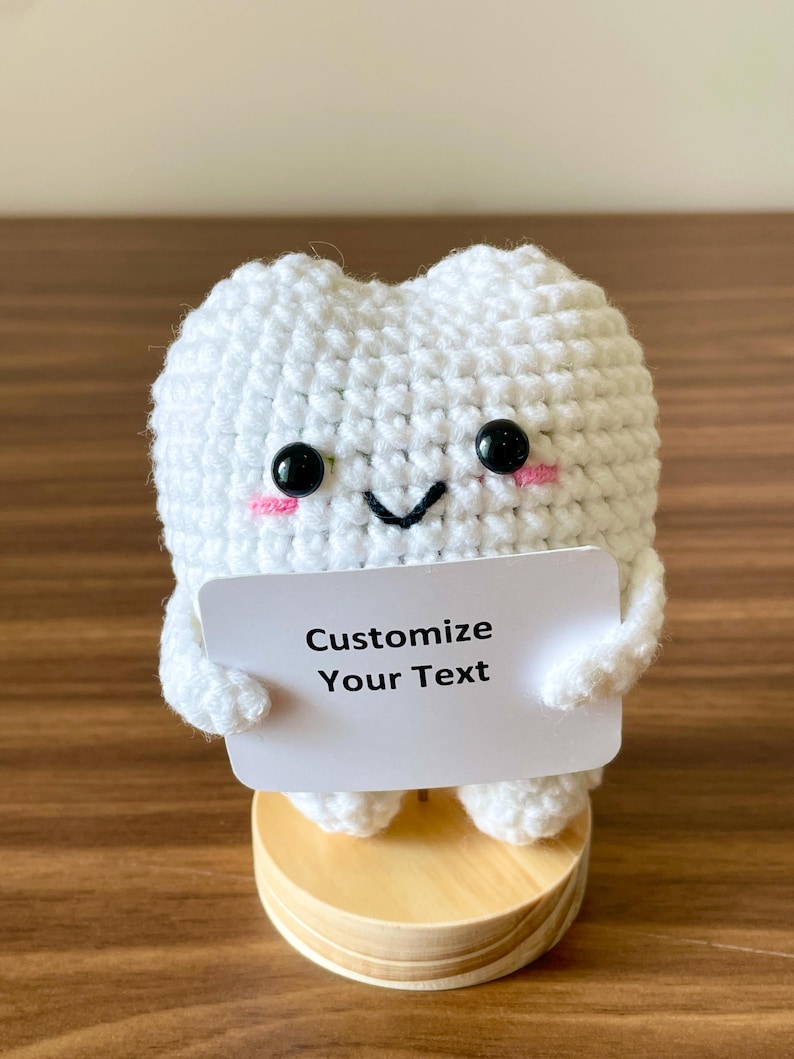May include: A handmade white crochet tooth with black button eyes, pink cheeks, and a smiling mouth. The tooth holds a small white sign that reads "Customize Your Text." It is mounted on a small wooden base.