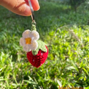 May include: A crocheted strawberry keyring with a white flower and green leaves attached to a silver key ring. The keyring is hanging from a hand.