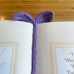 May include: A purple crocheted bookmark with two leaf-shaped ends. The bookmark is placed in a book with the text "May's house" visible.