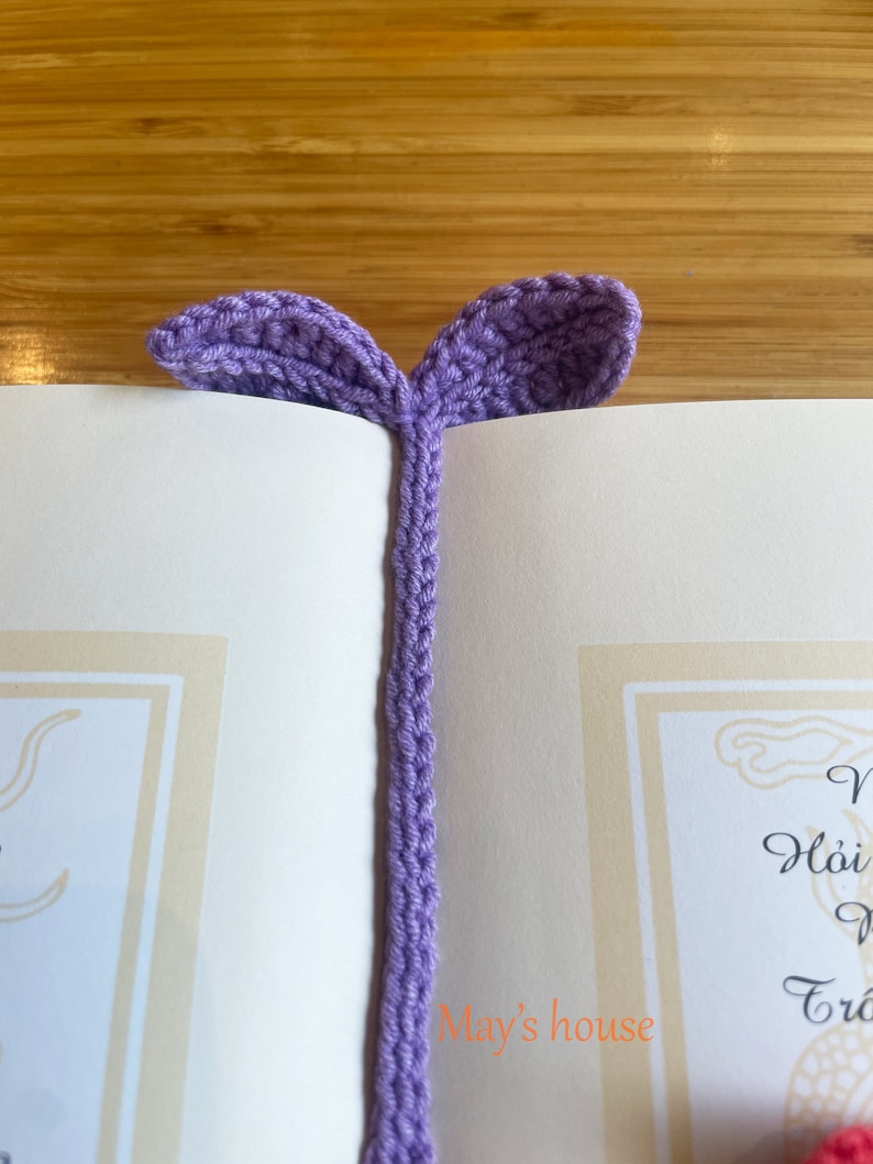 May include: A purple crocheted bookmark with two leaf-shaped ends. The bookmark is placed in a book with the text "May's house" visible.