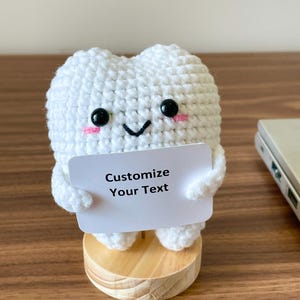May include: A handmade, white crochet tooth figure with black eyes and pink cheeks, holding a white sign that reads "Customize Your Text." The tooth is mounted on a small, round wooden base. The background is a wooden surface.
