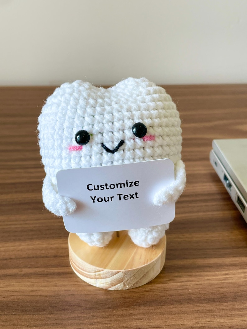 May include: A handmade, white crochet tooth figure with black eyes and pink cheeks, holding a white sign that reads "Customize Your Text." The tooth is mounted on a small, round wooden base. The background is a wooden surface.
