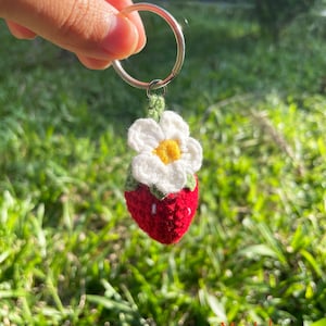 May include: A crocheted strawberry keyring with a white flower and green leaves. The keyring is attached to a silver ring.