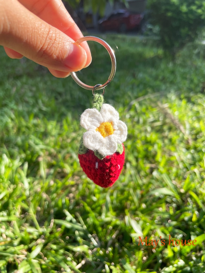 May include: A crocheted strawberry keyring with a white flower and green leaves. The keyring is attached to a silver ring.