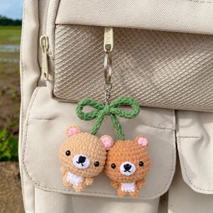 以下が含まれることがあります： 薄茶色と茶色の毛糸で編まれた2つの小さなクマの頭が付いたかぎ針編みのキーホルダー。それぞれのクマにはピンクの耳と白い口元があり、緑色の編み込みの茎が上部でリボン状に結ばれ、銀色の金属リングに取り付けられています。