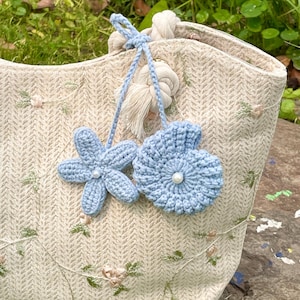 May include: A beige tote bag with a woven texture and floral embroidery. Two light blue crochet flower charms hang from the handle. One charm is a five-petal flower, and the other is a seashell shape with a pearl centre.
