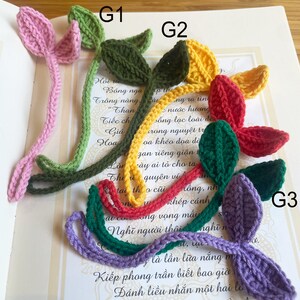 May include: Three crocheted bookmarks in the shape of leaves. The bookmarks are in pink, green, and purple. The bookmarks are on a white surface with text in a foreign language.