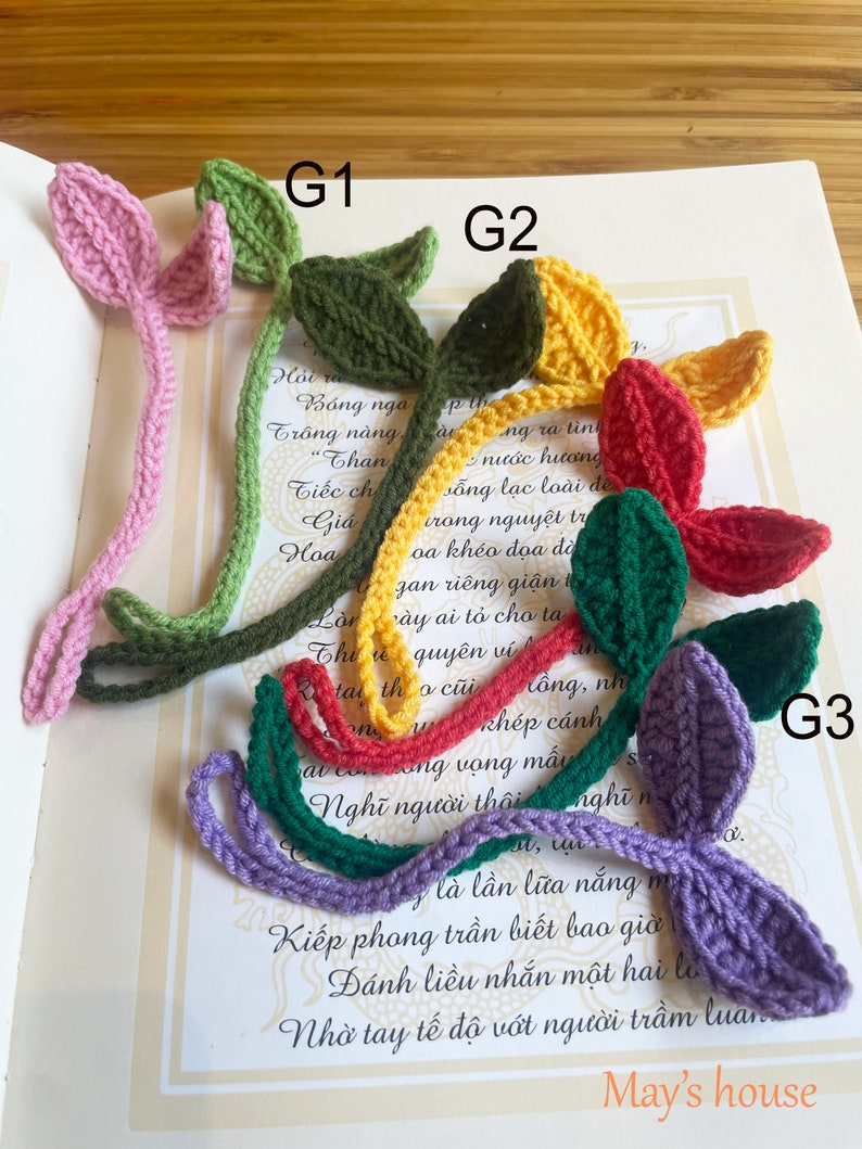 May include: Three crocheted bookmarks in the shape of leaves. The bookmarks are in pink, green, and purple. The bookmarks are on a white surface with text in a foreign language.