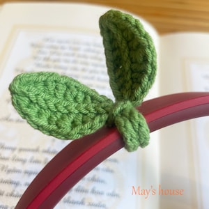 May include: Green crocheted leaf-shaped headphone charms. The charms are attached to a red headphone band. The text "May's house" is visible in the background.