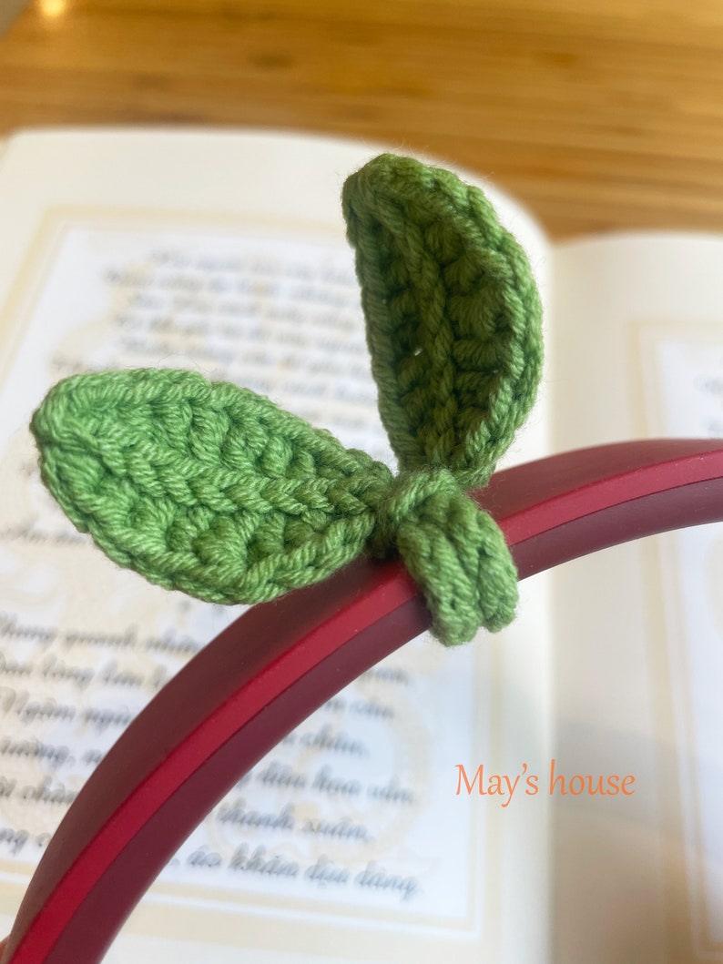 May include: Green crocheted leaf-shaped headphone charms. The charms are attached to a red headphone band. The text "May's house" is visible in the background.