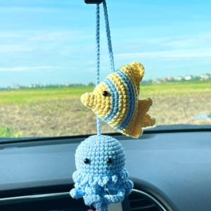 May include: A crochet car hanging ornament featuring a yellow and light blue striped fish and a light blue jellyfish with black bead eyes, suspended from a light blue yarn cord. The two marine creatures are connected by a single vertical string.