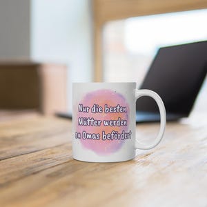 Könnte beinhalten: Weiße Keramik-Tasse mit Henkel, mit dem Text "Nur die besten Mütter werden zu Omas befördert" in Lila, vor einem rosa-violetten Hintergrund im Aquarellstil. Die Tasse steht auf einer Holzoberfläche.