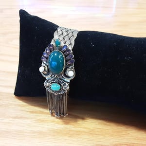 May include: A silver-toned braided cuff bracelet featuring a large oval dark green central stone, surrounded by smaller purple and white accents. Below the main setting, a smaller turquoise-colored stone sits above a dangling silver chain fringe.