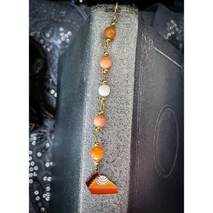 Puede incluir: Un marcapáginas dorado con un amuleto de pastel de calabaza y cuentas de color naranja, blanco y melocotón. El marcapáginas está colgado de un libro.