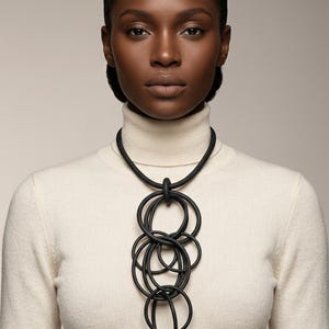 May include: A black statement necklace featuring multiple interlocking oval and circular shapes. The necklace is made of a matte black material and hangs from a black cord. The model is wearing a cream-colored turtleneck.