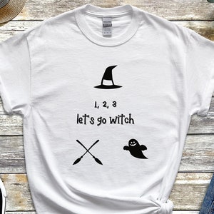 May include: White t-shirt with the text "1, 2, 3 let's go witch" and witch-themed graphics. The design includes a witch hat, crossed arrows, and a ghost. The shirt is paired with black and white sneakers and blue jeans.