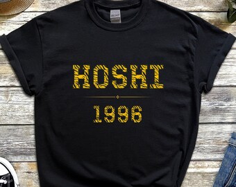 May include: Black t-shirt with the word "HOSHI" and the number "1996" in a yellow tiger stripe design. The shirt is displayed on a wooden surface with a pair of black and white sneakers, a straw hat, and denim shorts.