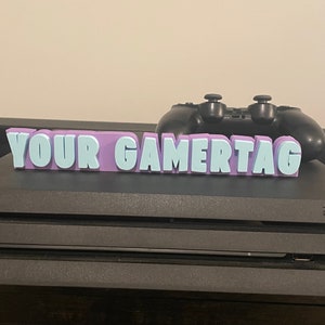 May include: A purple and white 3D printed sign that says "YOUR GAMERTAG" on top of a black Sony PlayStation 4 console.
