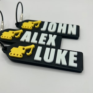 May include: Three black keychains with yellow construction vehicle graphics and white text. The names "JOHN", "ALEX", and "LUKE" are printed on the keychains.