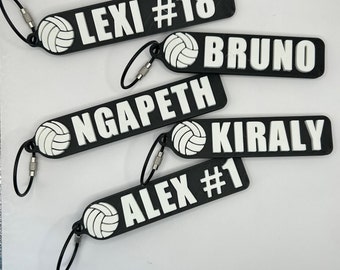 May include: Four black and white volleyball keychain tags with a volleyball graphic and names. The names are "LEXI #18", "BRUNO", "NGAPETH", and "KIRALY". One tag also has the name "ALEX #1".