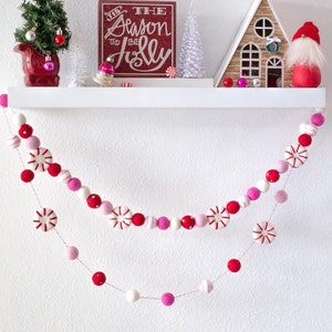 May include: A white shelf with a red and white felt garland with pink and white pom poms. The shelf is decorated with a small Christmas tree, a red sign that says "The Season to be Jolly", a small white Christmas tree, a small wooden house, and a red gnome.