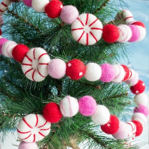May include: A festive garland featuring a mix of red, white, and pink felt balls and peppermint-shaped ornaments. The garland is draped over a small evergreen tree, creating a cheerful holiday decoration. The color palette includes shades of red, pink, and white.