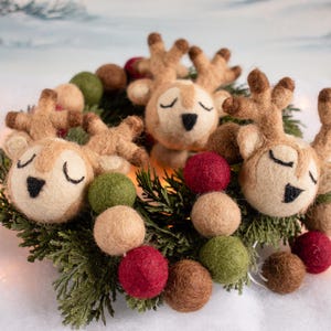 May include: A decorative wreath featuring three needle-felted reindeer heads with closed eyes and brown antlers, interspersed with small felt balls in shades of red, olive green, and tan, all nestled within a ring of green pine needles.
