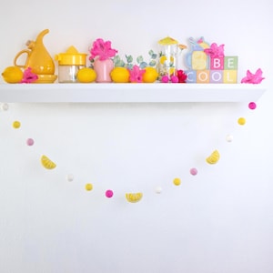 May include: A white shelf with a yellow pitcher, a yellow citrus squeezer, a pink flower, a glass jar, lemons, and a sign that says "Be Cool". A yellow and pink garland hangs below the shelf.