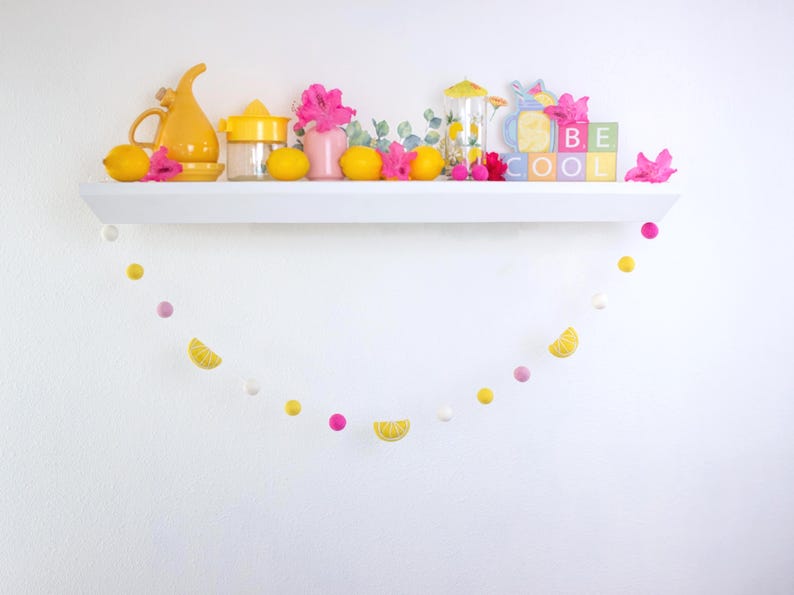 May include: A white shelf with a yellow pitcher, a yellow citrus squeezer, a pink flower, a glass jar, lemons, and a sign that says "Be Cool". A yellow and pink garland hangs below the shelf.