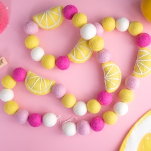 May include: A felt garland with yellow lemon slices and pink, white, and lavender pom poms. The garland is on a pink background. The word "Summer" is written on a wooden block.