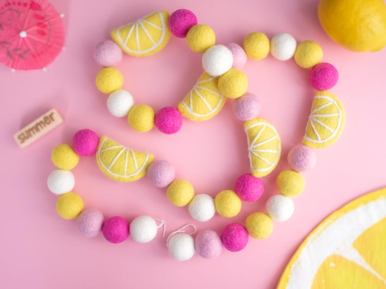 May include: A felt garland with yellow lemon slices and pink, white, and lavender pom poms. The garland is on a pink background. The word "Summer" is written on a wooden block.