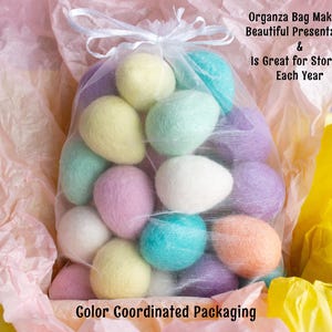 May include: A sheer organza bag filled with pastel coloured felt Easter eggs. The bag is sitting on a pink and yellow tissue paper background. The text "Organza Bag Makes a Beautiful Presentation & Is Great for Storage Each Year" is written above the bag. The text "Color Coordinated Packaging" is written below the bag.