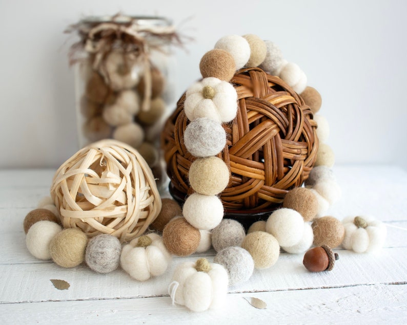 May include: A garland of white, beige, and brown felt balls with a woven wicker ball in the center. The garland is on a white surface with a small brown acorn.